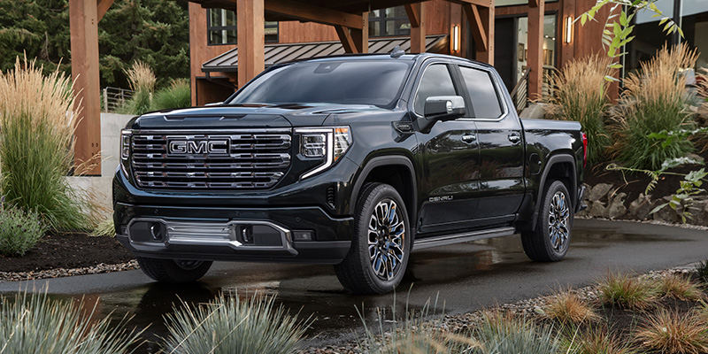 A shining black 2026 GMC Sierra 1500 is parked on a nice asphalt carport beside a beautiful modern desert home or office and its garden.