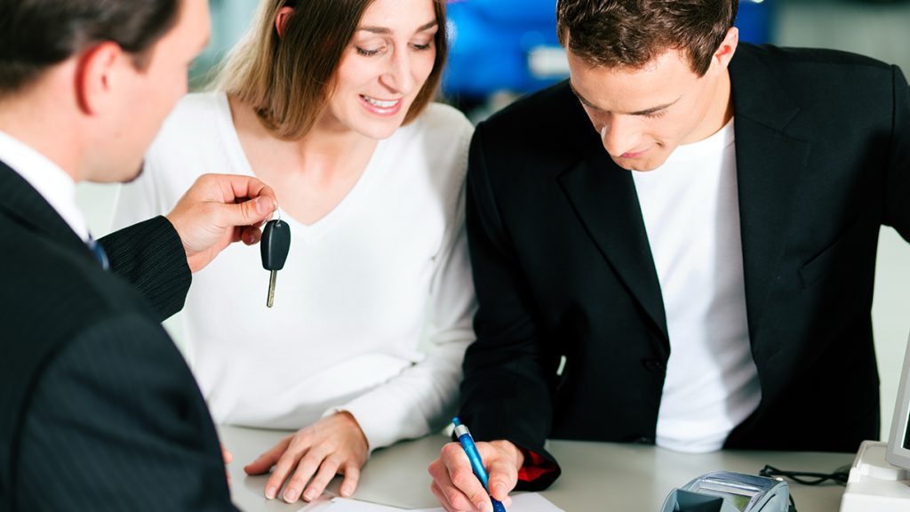 A smiling couple sign New Car Financing paperwork as the sales representative hands them the key to the vehicle.